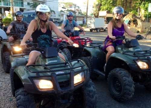 Woman in a white helmet and sunglasses on a green ATV with other riders behind her on a village street