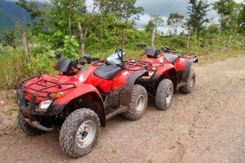 Two muddy red ATVs parked on a gravel road with green hills behind