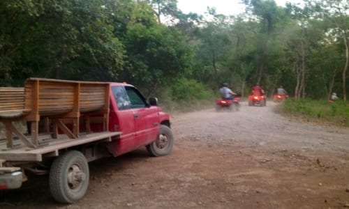 Red pickup truck with a wooden bed on a dirt road as ATV riders drive ahead