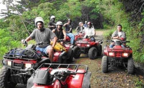Group of riders on red ATVs on a dirt trail through green forest, some wearing helmets