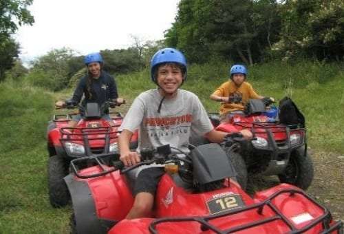 Smiling boy in a blue helmet on a red ATV with two other riders on a grassy track