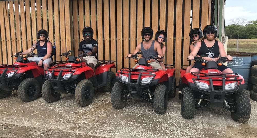 Six smiling riders in helmets and sunglasses on red ATVs in front of a wooden fence