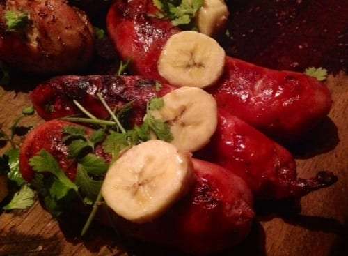 Grilled red sausages topped with banana slices and fresh cilantro on a wooden board