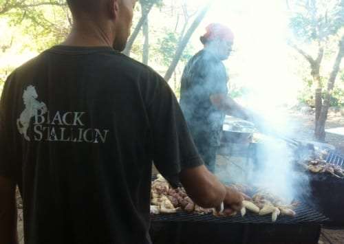 Man in a Black Stallion shirt grilling sausages over smoke while a cook works behind him