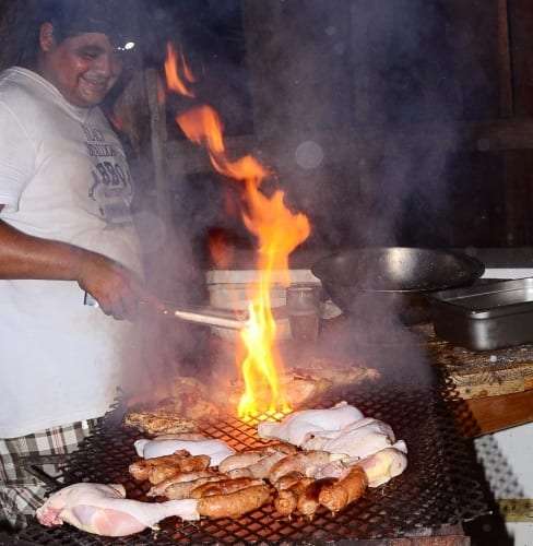 Smiling cook tending chicken and sausages over a flaming charcoal grill