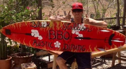 Man in a red cap holding a surfboard painted with Black Stallion BBQ Rustic Dining