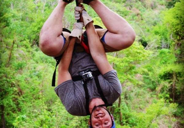 Smiling rider hanging upside down in a harness on a zip line above green forest