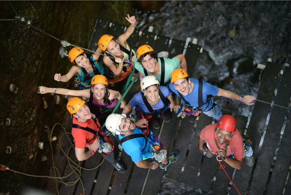 Group of smiling people in orange helmets and harnesses on a wooden platform, seen from above