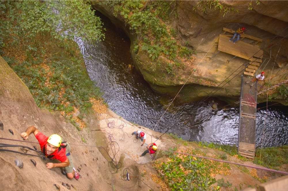 Climber in a yellow helmet scaling a rock wall above a river gorge with guides and a hanging bridge