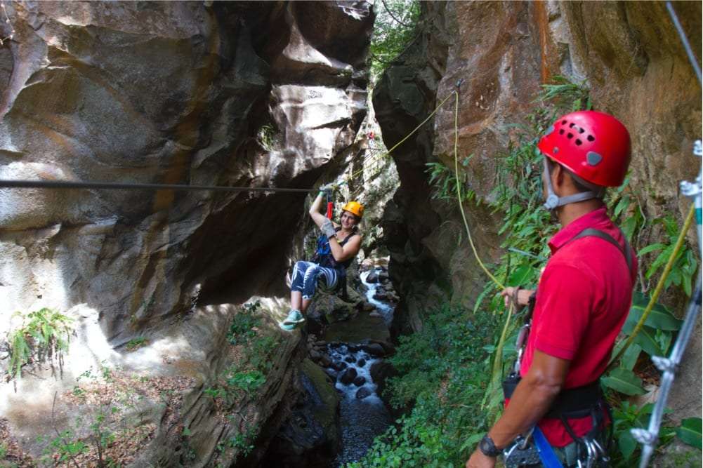 Woman in an orange helmet zip lining across a narrow rock canyon while a guide in a red helmet watches