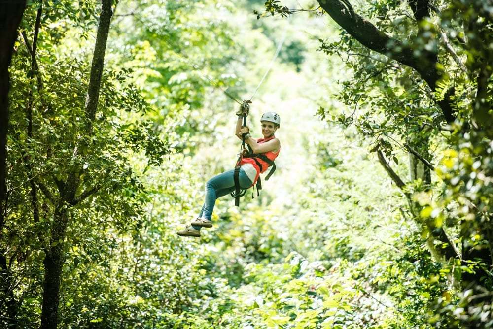 Smiling woman in a white helmet riding a zip line through dense green forest