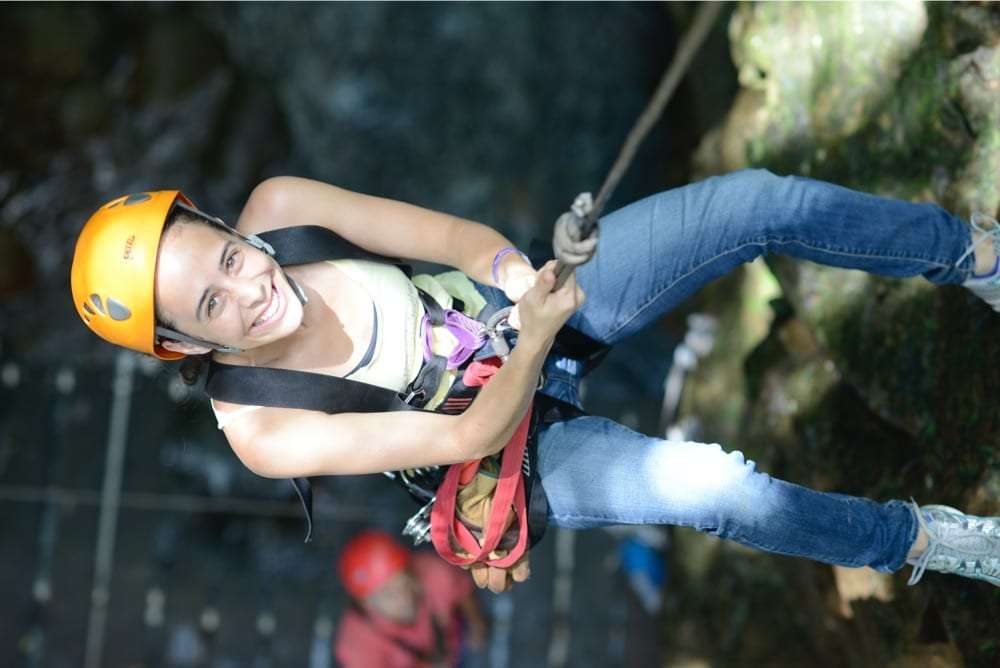Woman in an orange helmet and harness smiling as she hangs from a rope on a rappel