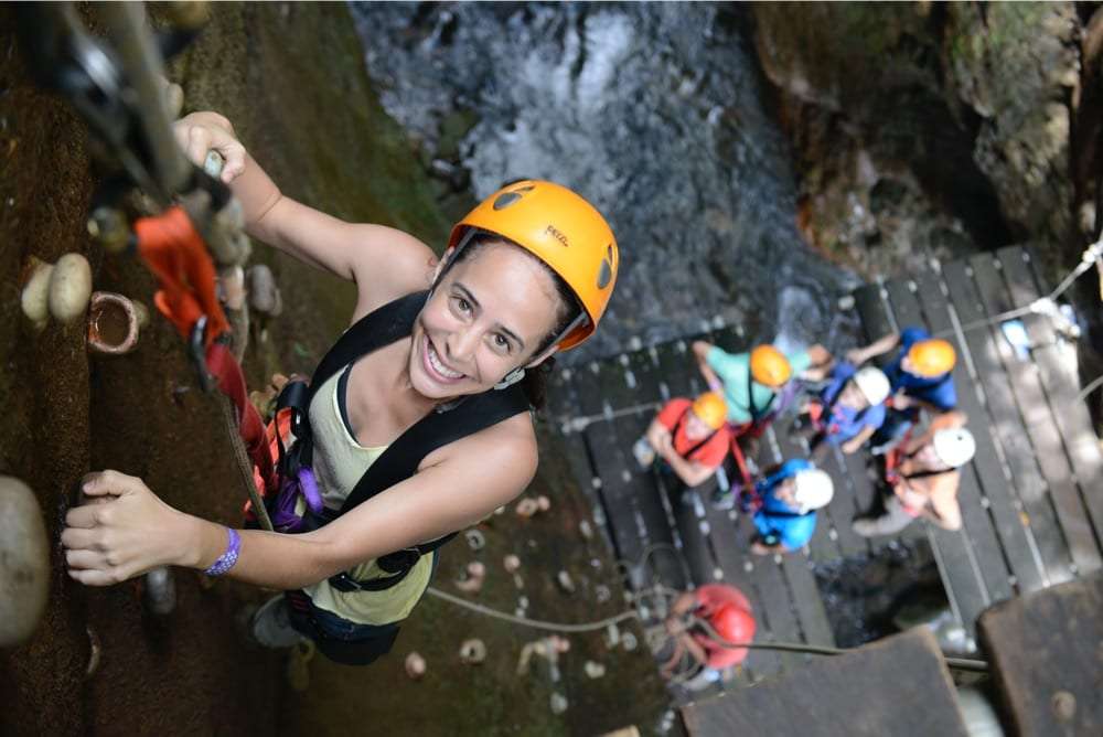 Woman in an orange helmet climbing a wall while guests wait on a wooden platform below