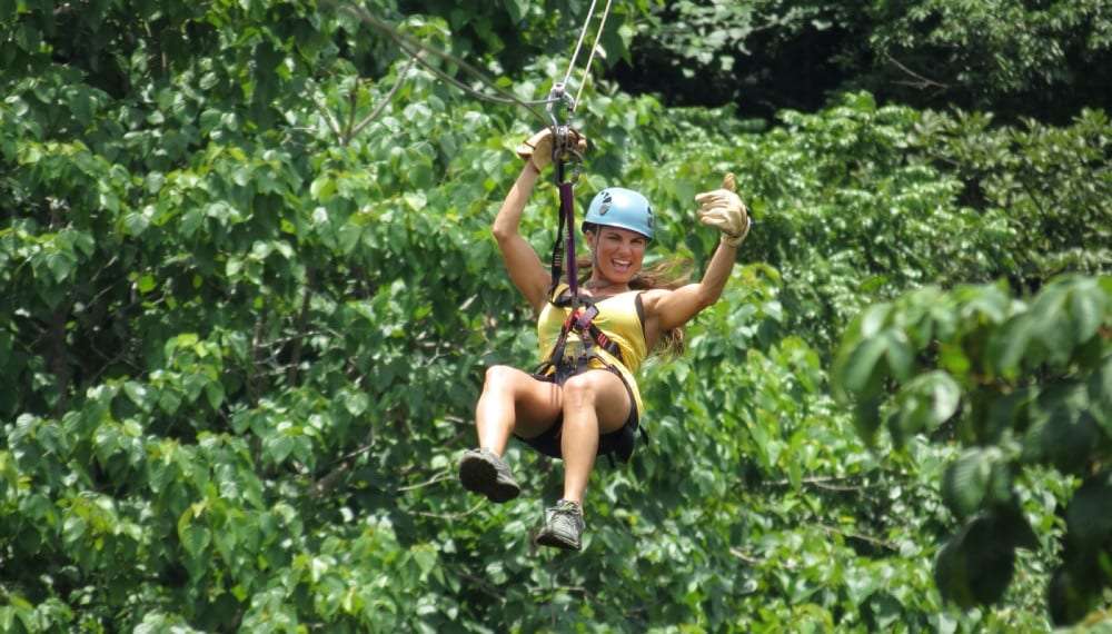 Smiling woman in a blue helmet giving a thumbs up on a zip line above the tree canopy