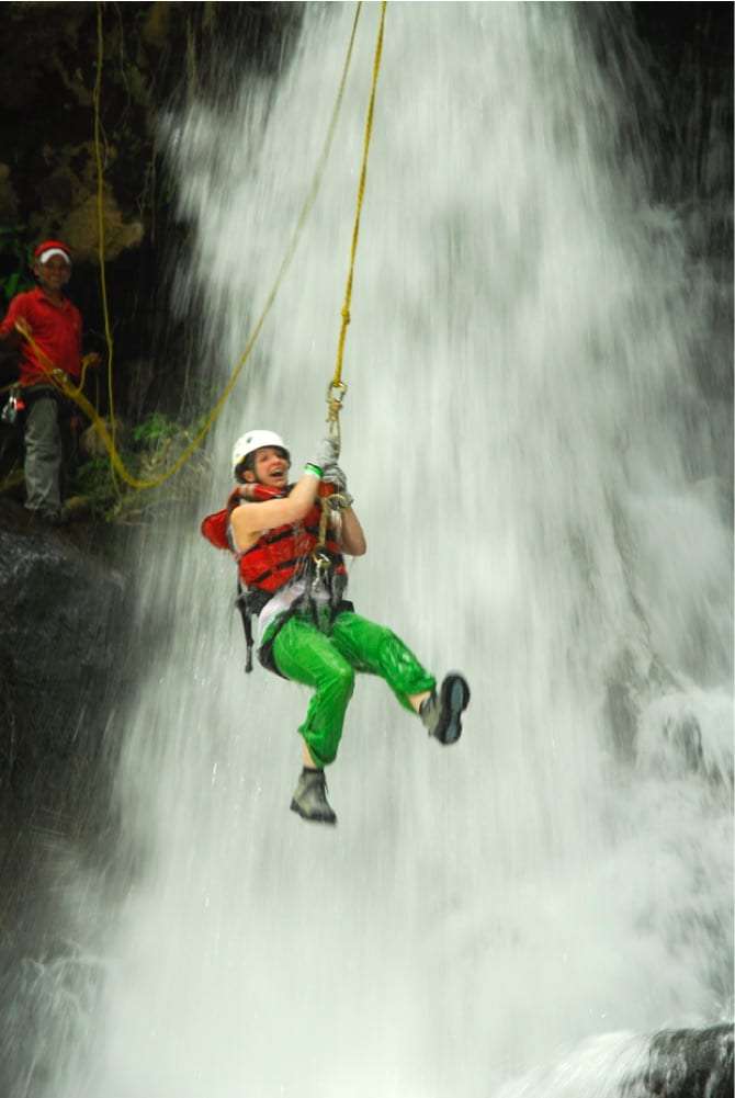 Woman in a life vest and green waterproof pants rappelling through a waterfall