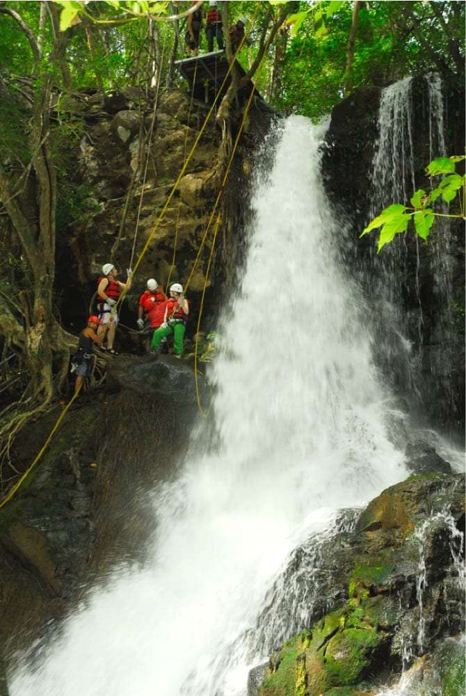 Guides and guests in helmets on a rock ledge beside a waterfall with a platform above