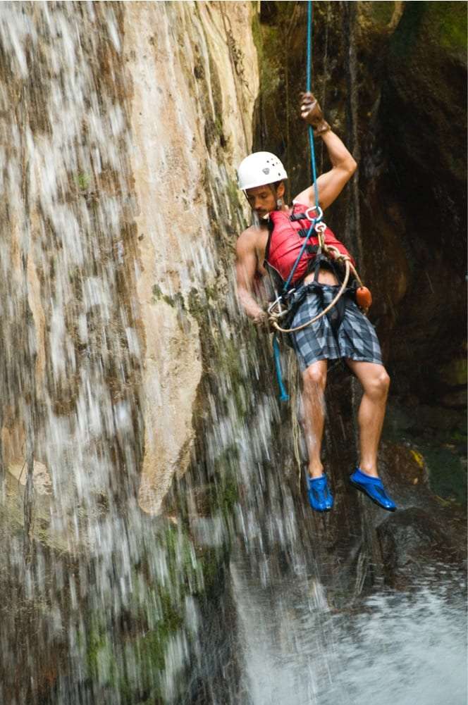 Man in a white helmet and life vest rappelling down a wet rock beside a waterfall