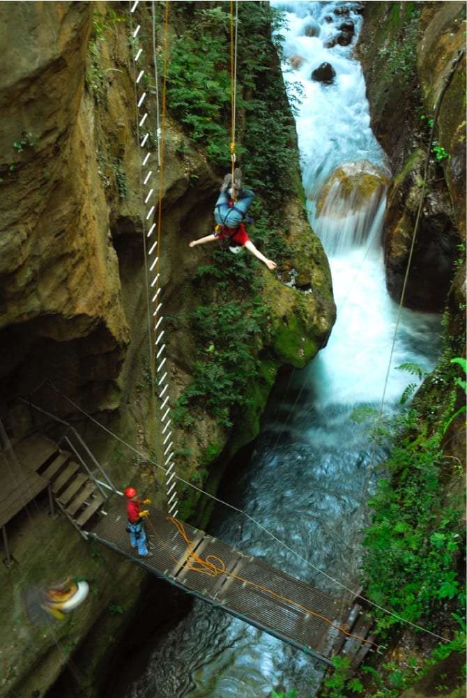 Guest hanging upside down on a zip line above a narrow gorge, with a guide on a metal walkway below