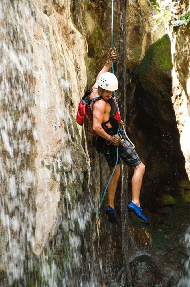 Shirtless man in a white helmet rappelling down a wet rock wall on a blue rope