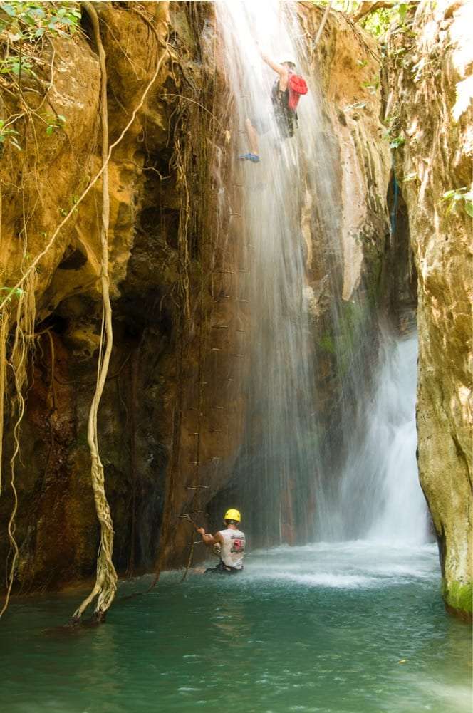 Climber descending a waterfall while a man in a yellow helmet waits in the pool below