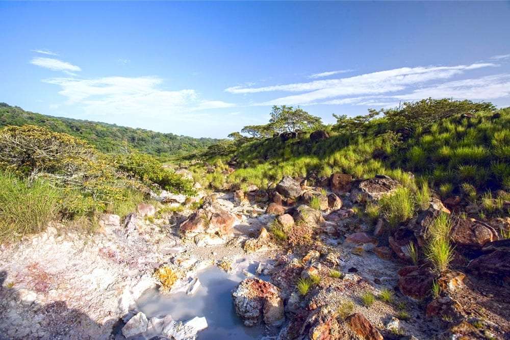 Small pool of milky water among rocks and grass on a volcanic hillside under blue sky