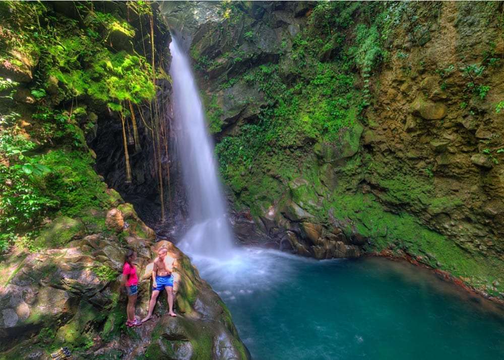 Two people standing on mossy rocks beside a waterfall and a turquoise pool