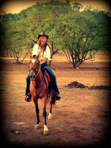 Laughing woman in a cowboy hat riding a chestnut horse along a dirt path