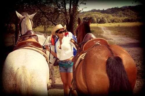 Woman in a straw hat and sunglasses standing between two saddled horses on a dirt road