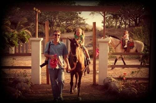 Man with a camera leading a horse carrying a young girl through a wooden gate