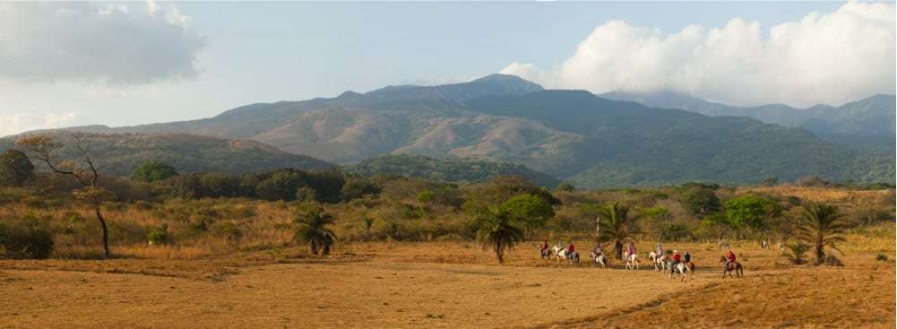 Small group of riders on horseback crossing dry grassland with mountains behind them
