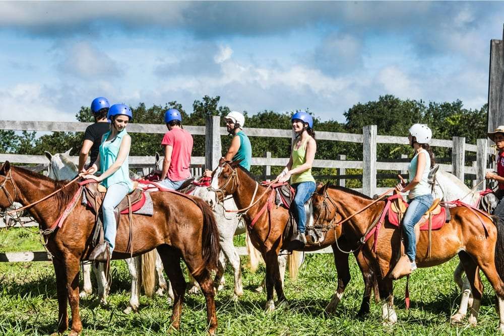 Riders in helmets sitting on horses beside a white wooden fence under a cloudy blue sky
