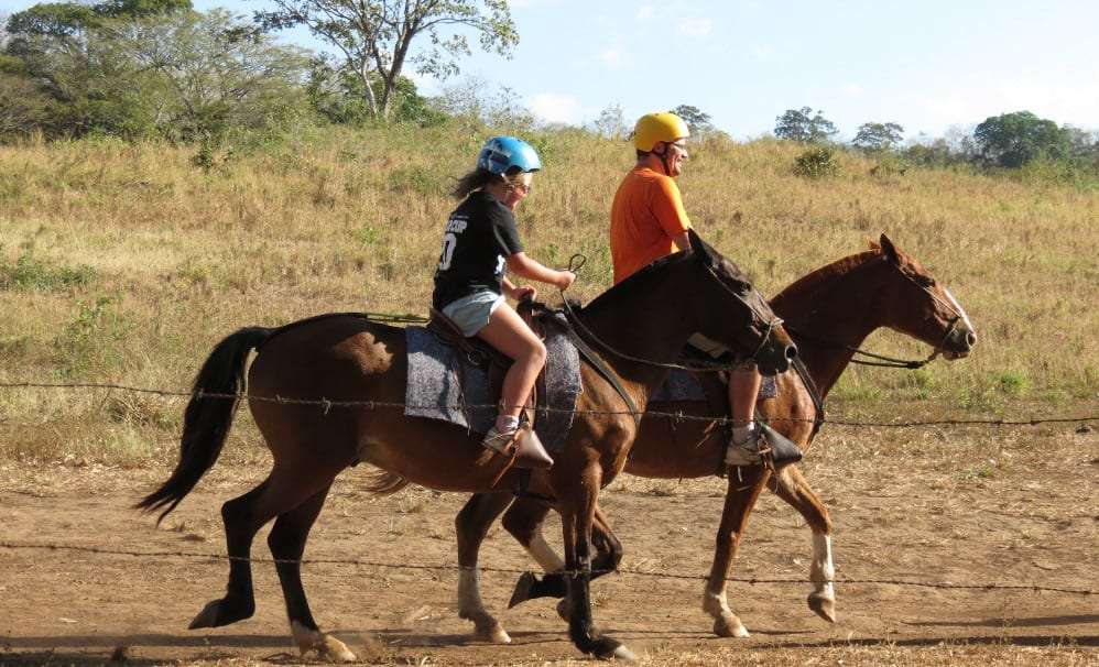 Two riders in helmets on brown horses beside a dry grass field
