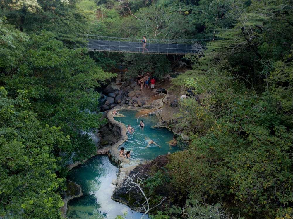 Aerial view of hot spring pools in a forest below a suspension bridge