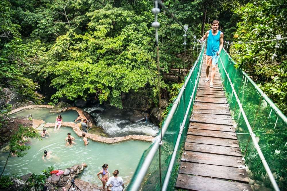 Man in a teal tank top walking across a green-railed suspension bridge above thermal pools with bathers