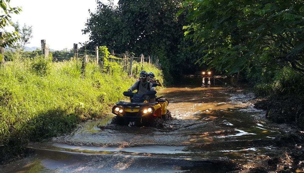 Two riders in helmets on a yellow ATV driving through a muddy stream