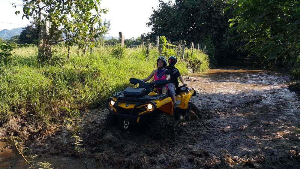 Two riders on a yellow ATV splashing through mud beside a grassy field