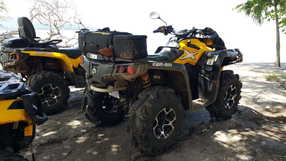 Mud-splattered yellow Can-Am ATV parked on a shaded sandy beach beside other ATVs