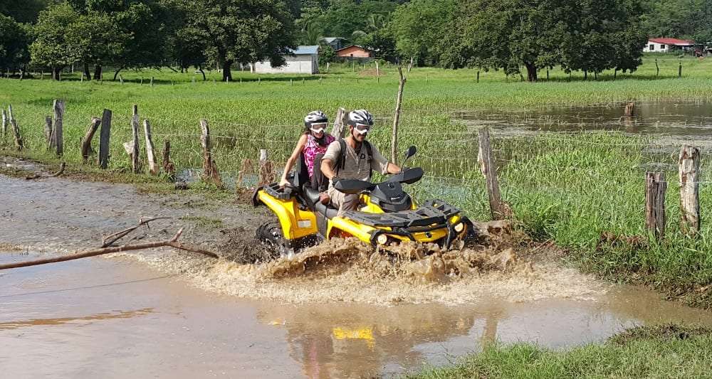 Two riders in helmets on a yellow ATV splashing through a flooded field with a farm behind