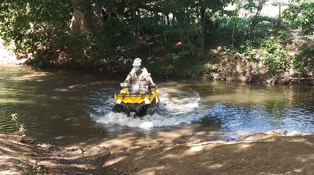 Rider in a white helmet driving a yellow ATV through a shallow river crossing