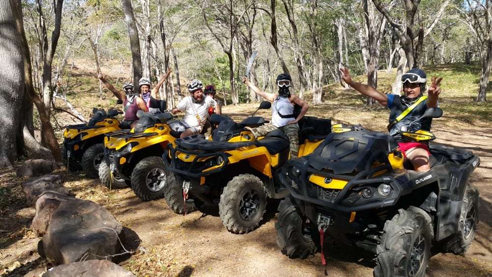 Group of ATV riders in helmets and goggles waving from yellow ATVs on a dry forest trail