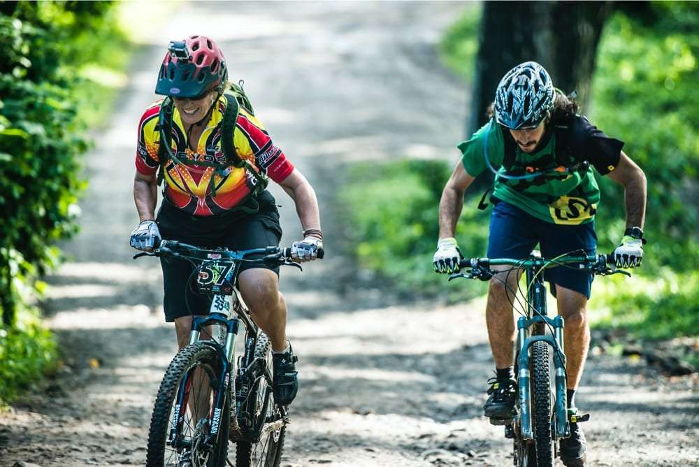 Two mountain bikers in helmets riding uphill on a shaded dirt road