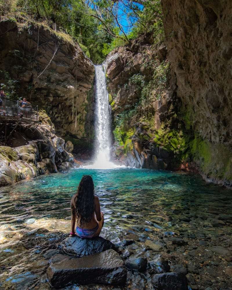 Woman sitting on a rock facing a tall waterfall and turquoise pool