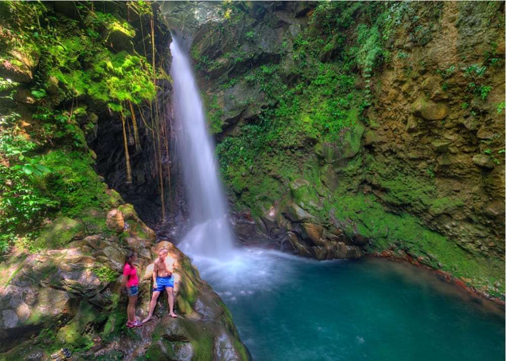 Two people standing on mossy rocks beside a waterfall and turquoise pool