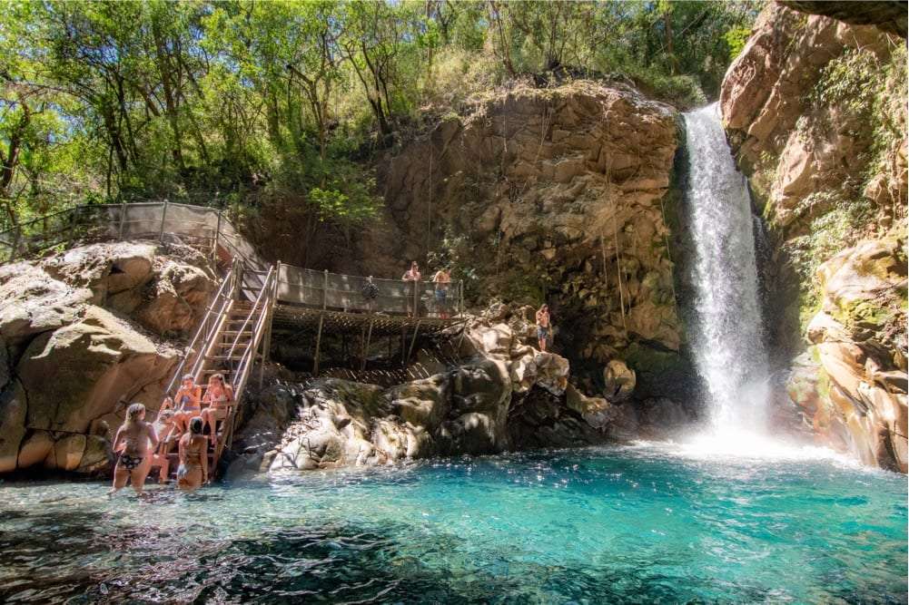 Swimmers in a turquoise pool below a waterfall with a wooden staircase and viewing deck