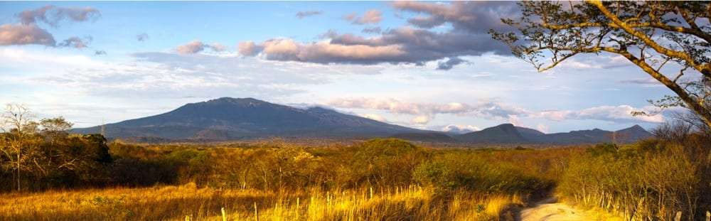 Volcano under a cloudy sky seen across dry grassland at golden hour