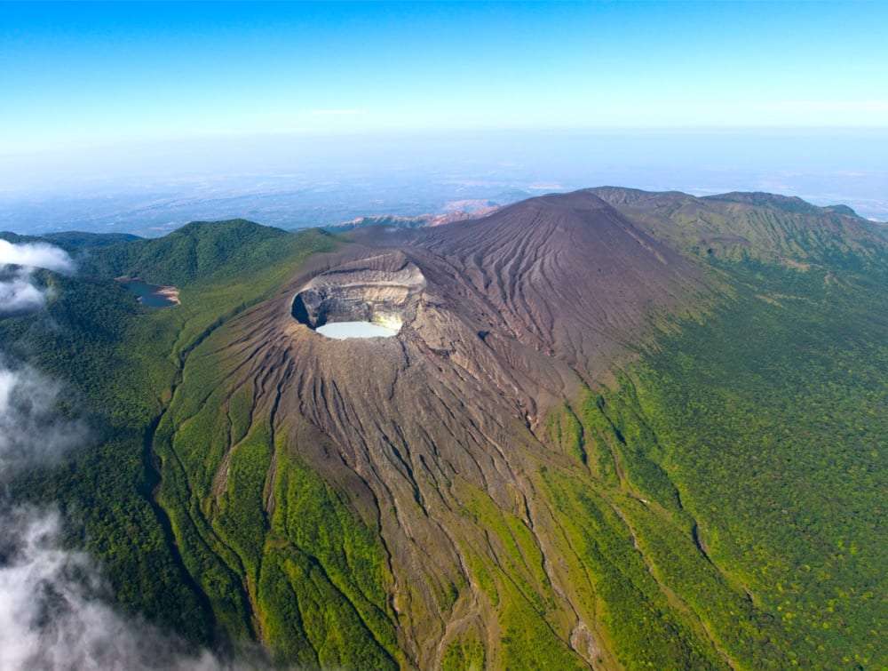 Aerial view of a volcano crater with a pale lake inside, surrounded by green slopes