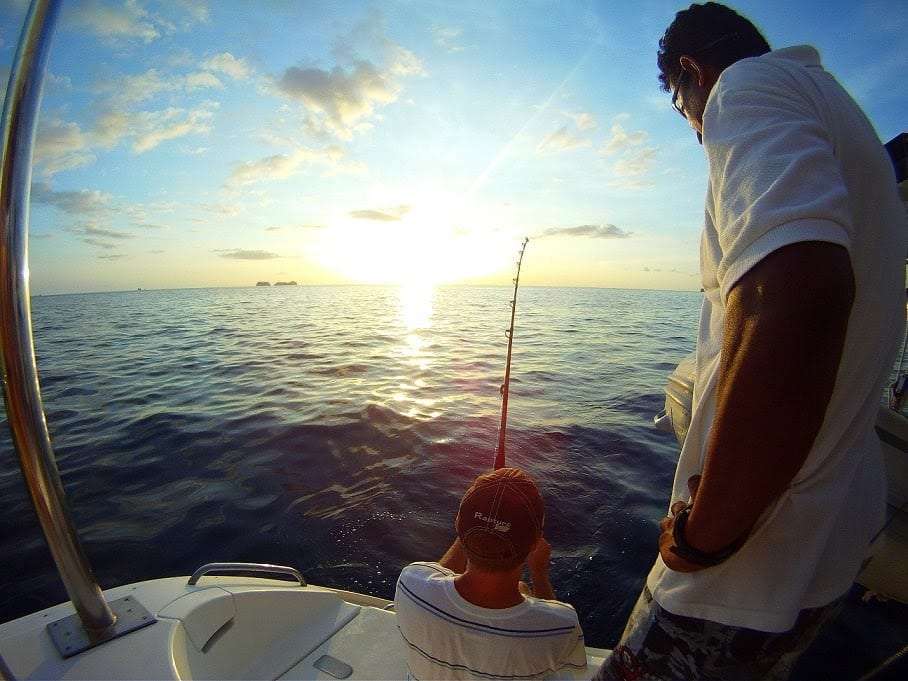 Two men on the back of a boat, one fishing, as the sun sets over the sea