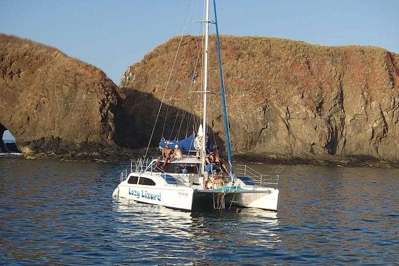 Passengers relaxing on the Lazy Lizard catamaran beside a rocky island with an arch