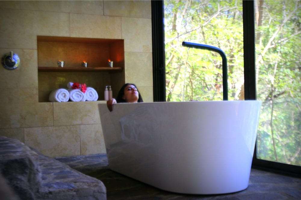 Woman relaxing in a white freestanding bathtub beside a window facing trees