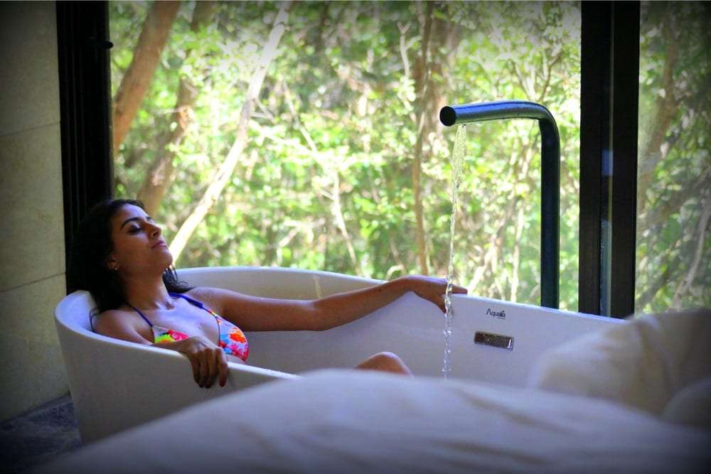 Woman relaxing in a white bathtub with a forest view as water pours from a spout